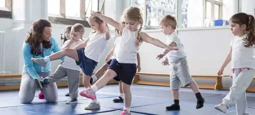 Ernährungs- und Bewegungspädagoge für Kinder | Kindergärtnerin hilft einem ihrer Schüler während einer Sportunterrichtsstunde.